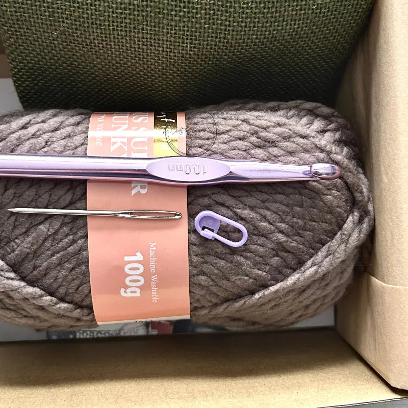 The yarn used in this crochet kit is lying in a box with a crochet hook, stitch marker and a tapestry needle.