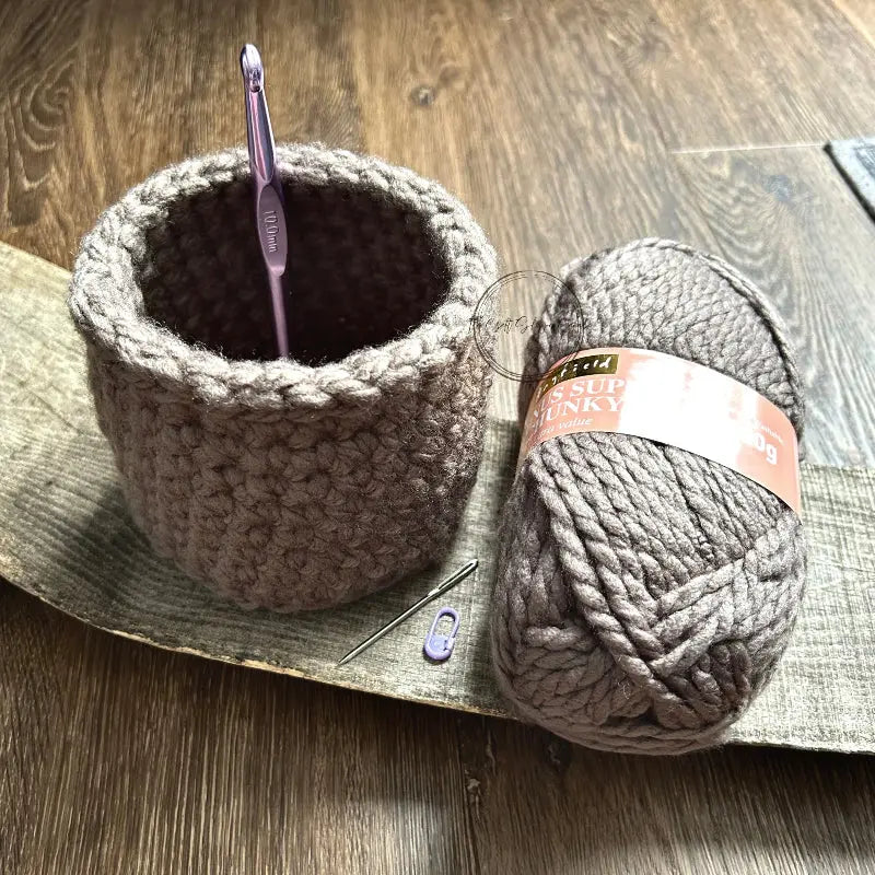 The storage basket, from this crochet kit, is sitting on a wooden board with a ball of yarn, crochet hook, tapestry needle and stitch marker.