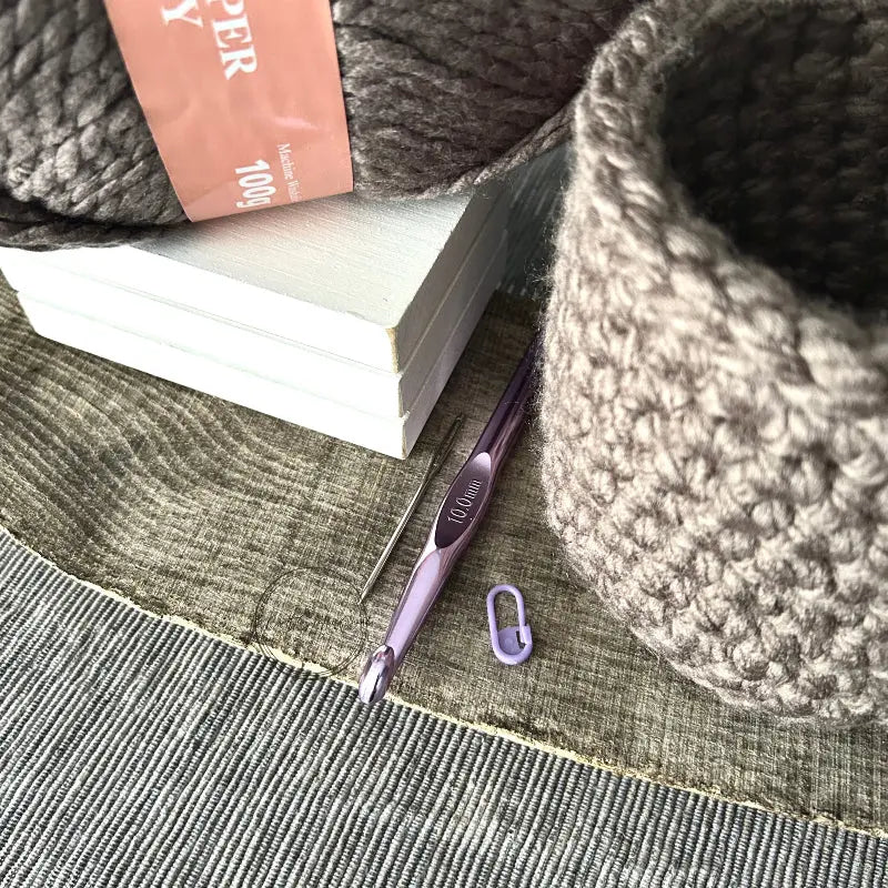 The storage basket, from this crochet kit, is sitting on a wooden board with a ball of yarn, crochet hook, tapestry needle and stitch marker.