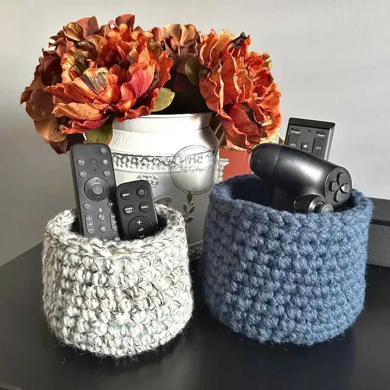 The storage baskets, from this crochet pattern, are sitting on a wooden table with TV and game remotes in the baskets.