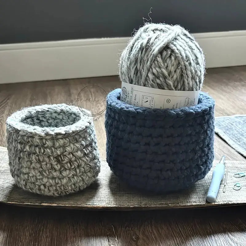 The storage baskets, from this crochet pattern, are sitting on a wooden board with a ball of yarn, a crochet hook and stitch markers beside them.