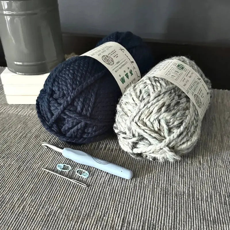 Two balls of yarn, used in this crochet pattern, are sitting on a table with a crochet hook, tapestry needle and stitch markers.