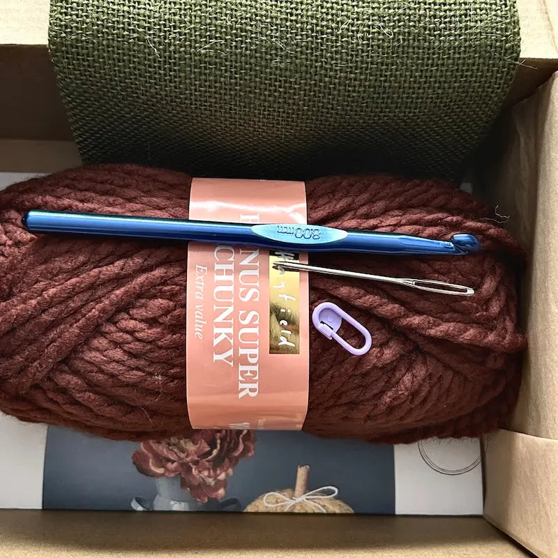 The yarn used in this crochet pattern is lying in a box with a crochet hook, stitch marker and a tapestry needle.