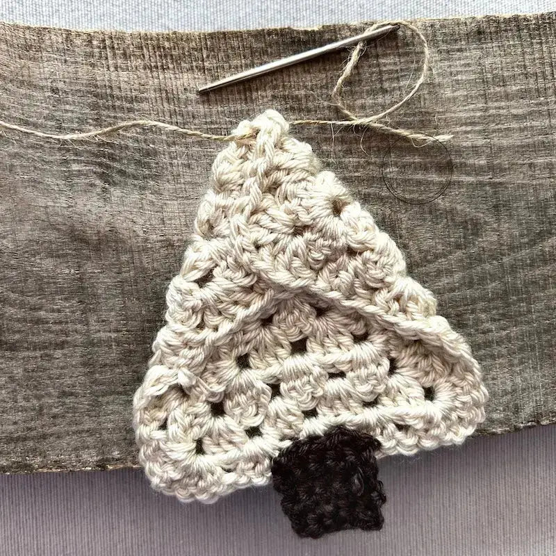 A tree ornament, made from this crochet kit, is sitting on a wooden board. The back of the ornament is facing you with a tapestry needle threading some twine through the top.