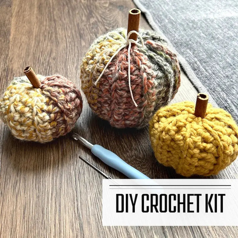 A crochet kit for a set of autumn pumpkins. The finished pumpkins are sitting on the floor with a crochet hook and tapestry needle in front of them.
