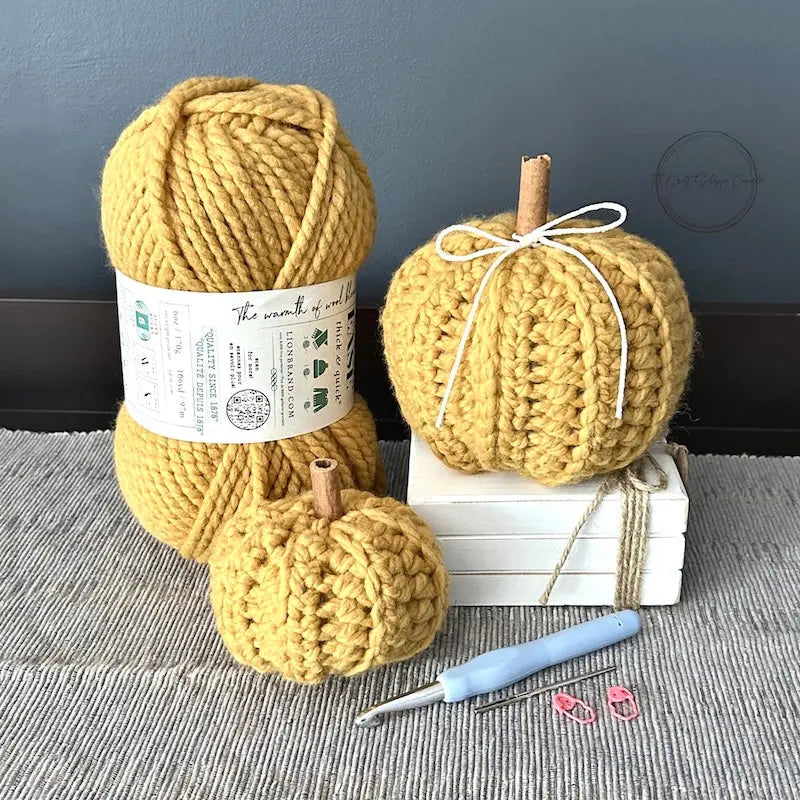 2 autumn pumpkins, made from this crochet kit are sitting on a buffet with a ball of yarn, crochet hook, a tapestry needle and stitch markers.