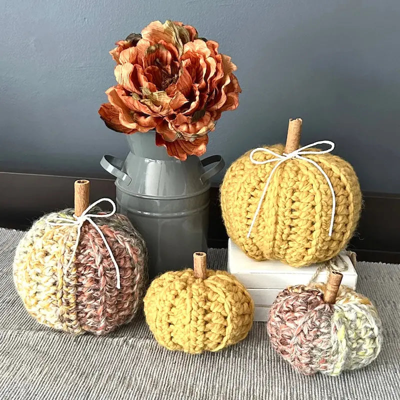 4 autumn pumpkins, made from this crochet pattern are sitting on a buffet with a vase of fall flowers beside them.