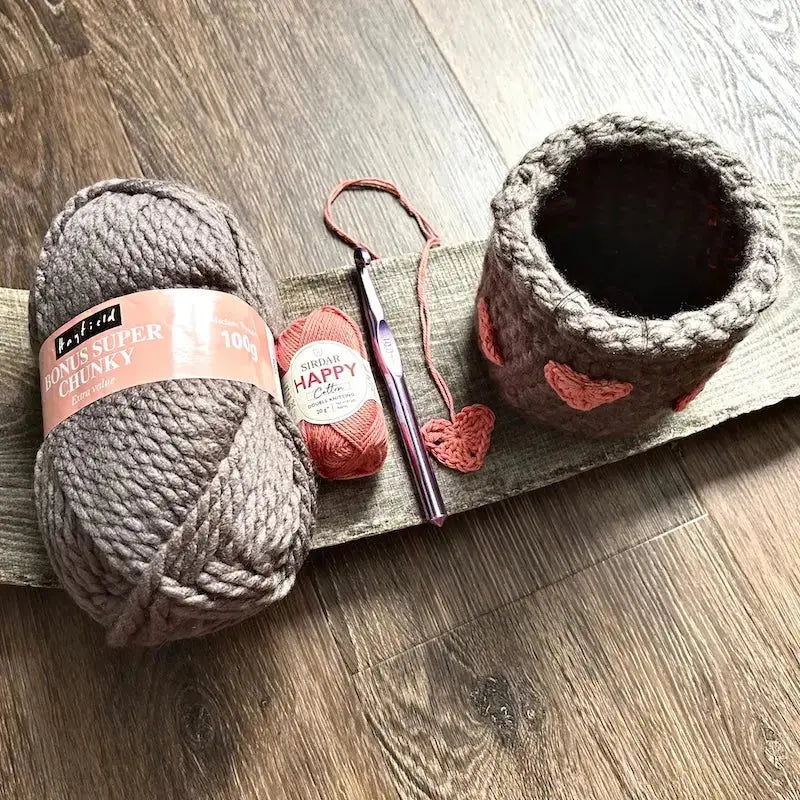The yarn, a crochet tool and the finished heart basket, made from this crochet pattern is sitting on a wooden board.
