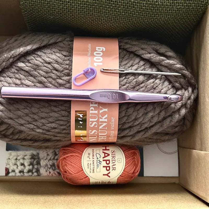 The yarn used in this crochet pattern is lying in a box with a crochet hook, stitch marker and a tapestry needle.