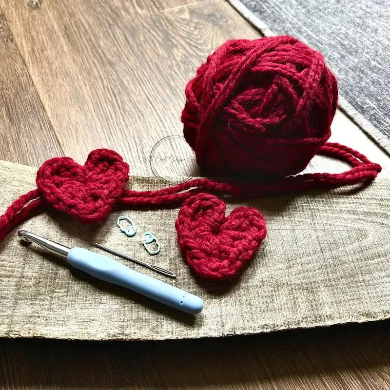 There are 2 hearts, sitting on a wooden board with red yarn and a crochet hook.