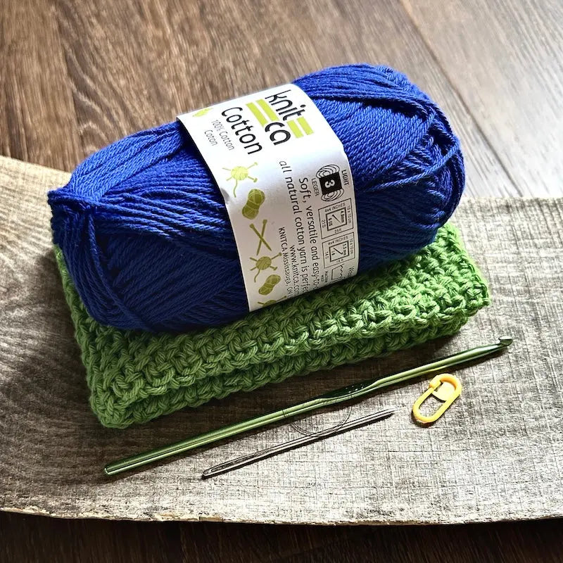 The finished washcloth, made from this crochet kit, is sitting on a wooden board with yarn, crochet hook, a tapestry needle and stitch marker.