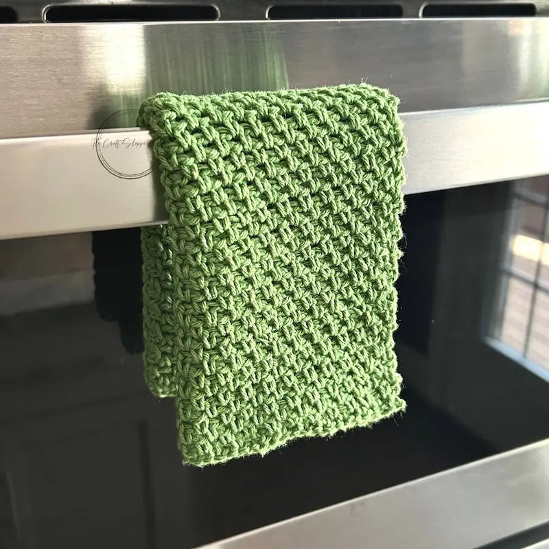 The finished washcloth, made from this crochet kit, is hanging off of a stove railing.