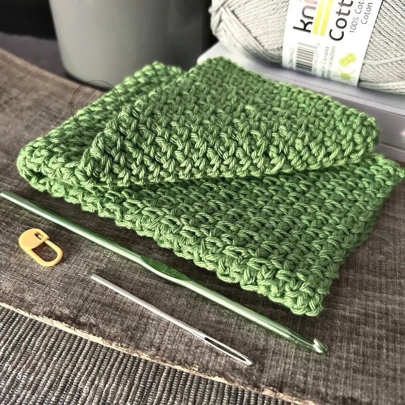The finished washcloth, made from this crochet kit, is sitting on a wooden board with yarn, crochet hook, a tapestry needle and stitch marker.