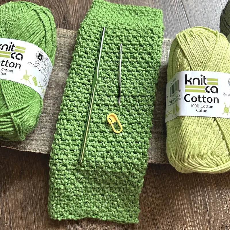 The finished washcloth, made from this crochet kit, is sitting on a wooden board with yarn, crochet hook, a tapestry needle and stitch marker.