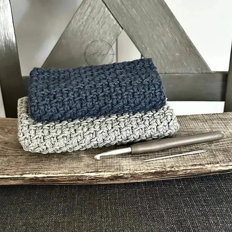 The finished washcloths, made from this crochet kit, are lying on a wooden board with a crochet hook.