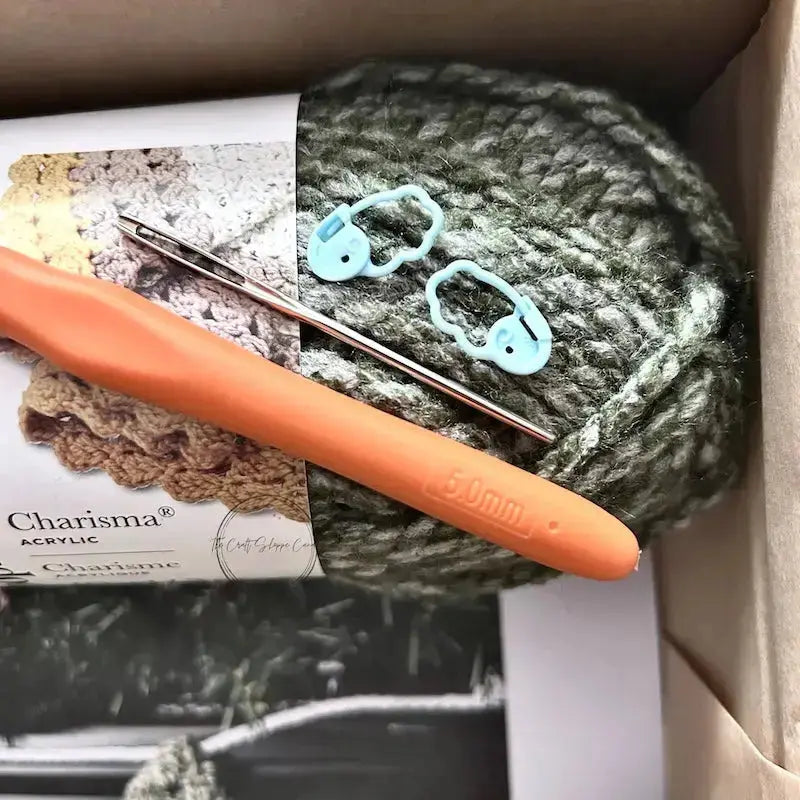 A ball of green yarn is sitting in a box with a crochet hook, a tapestry needle and 2 stitch markers.