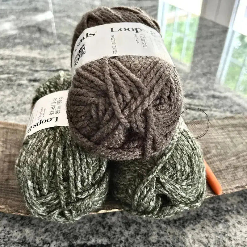 The yarn, that would be used with this crochet pattern, is sitting on a wooden board with a crochet hook.