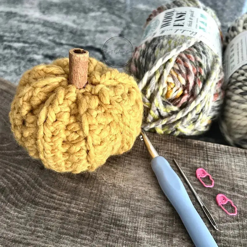A small pumpkin, made from the crochet kit, in sitting on a wooden board with a crochet hook and yarn.