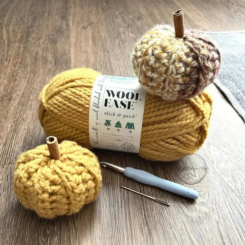 2 small crochet pumpkins, made from the crochet kit, are sitting on a ball of yarn and on the floor with a crochet hook in between them.