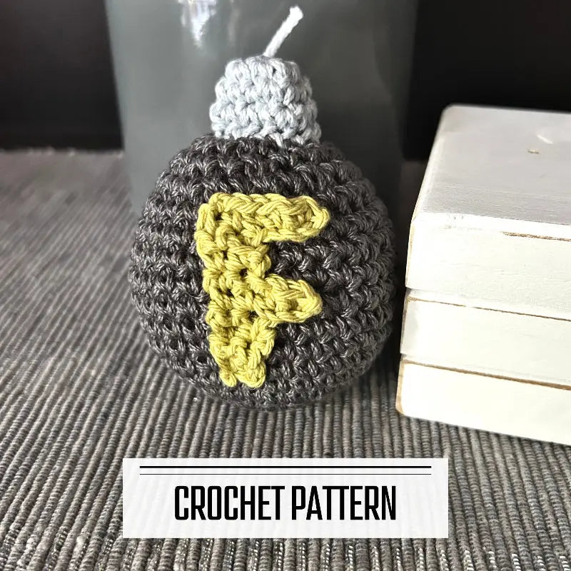 A DIY crochet pattern for an amigurumi plush F bomb. The F bomb is sitting on a table beside a wooden block.