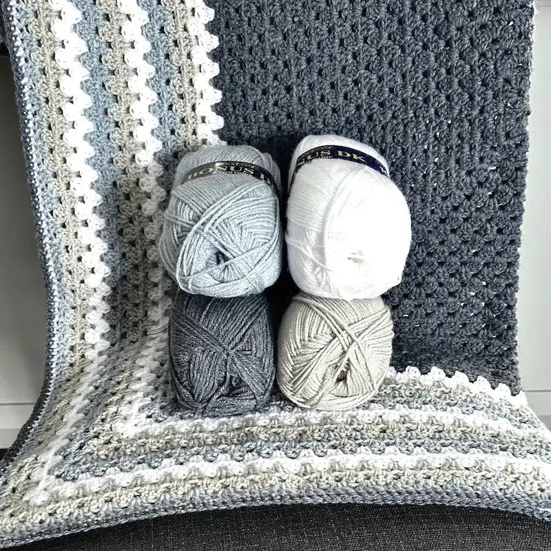 The finished granny square blanket, made from this crochet pattern, is lying over a chair with 4 balls of yarn.