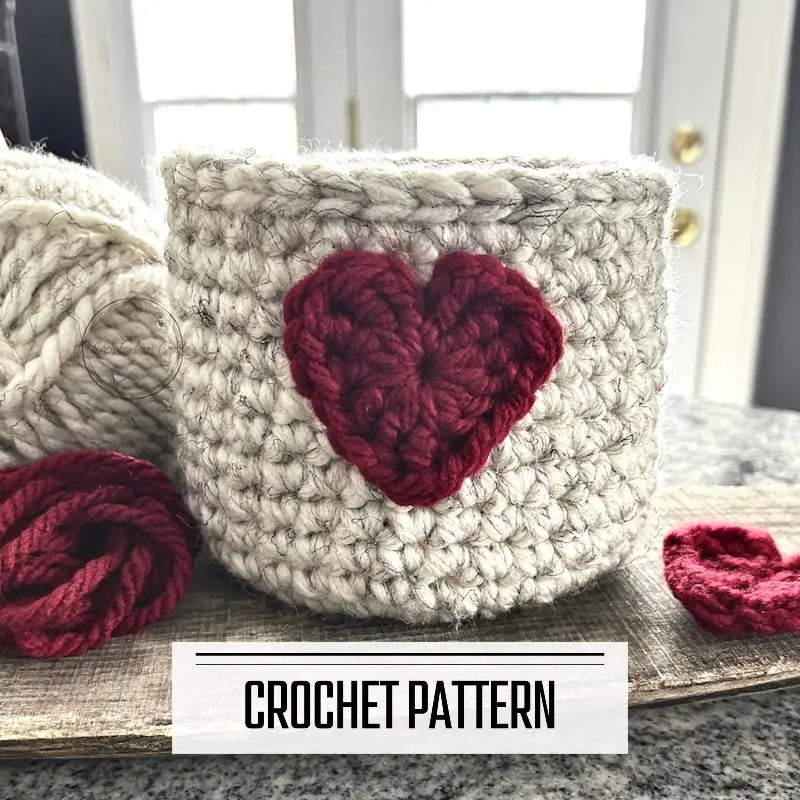 A digital PDF crochet pattern for a Valentine heart basket. The basket is sitting on a wooden board with yarn and a red heart beside it.