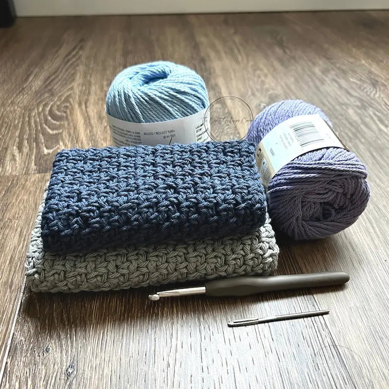 The finished washcloths, made from this crochet pattern, are sitting on the floor with yarn and a crochet hook.