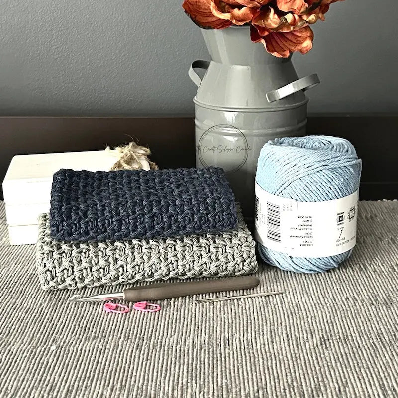 The finished washcloths, made from this crochet pattern, are lying on a table with yarn, a tapestry needle and a crochet hook.
