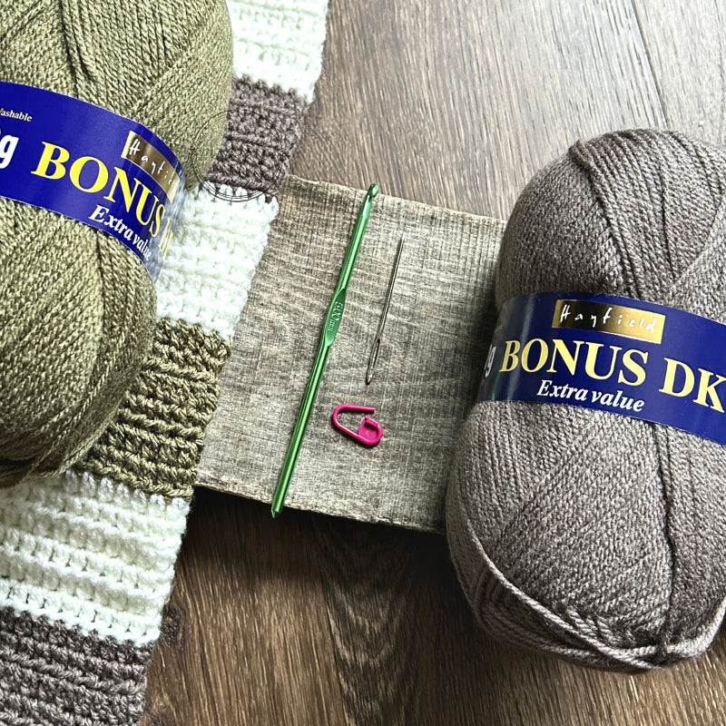 The tools needed to put the baby blanket together are lying on a wooden board. There are 2 balls of yarn, a crochet hook, tapestry needle and stitch marker.