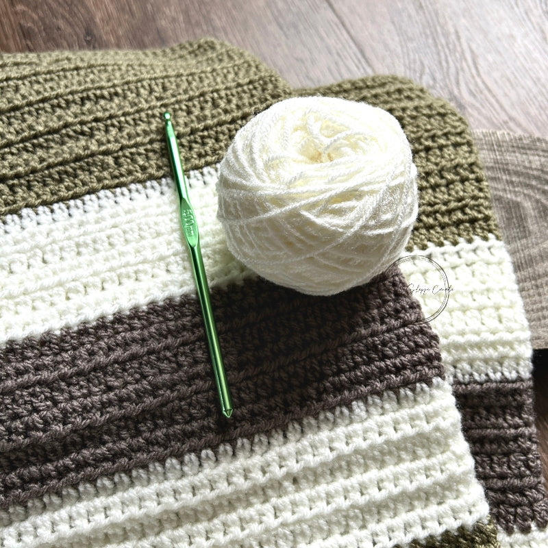 The finished baby blanket, made from this crochet kit, is lying on a wooden board with a ball of yarn and a crochet hook.