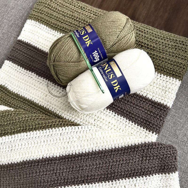 The finished baby blanket, made from this crochet kit, is draped over a couch with 2 balls of yarn and a crochet hook.