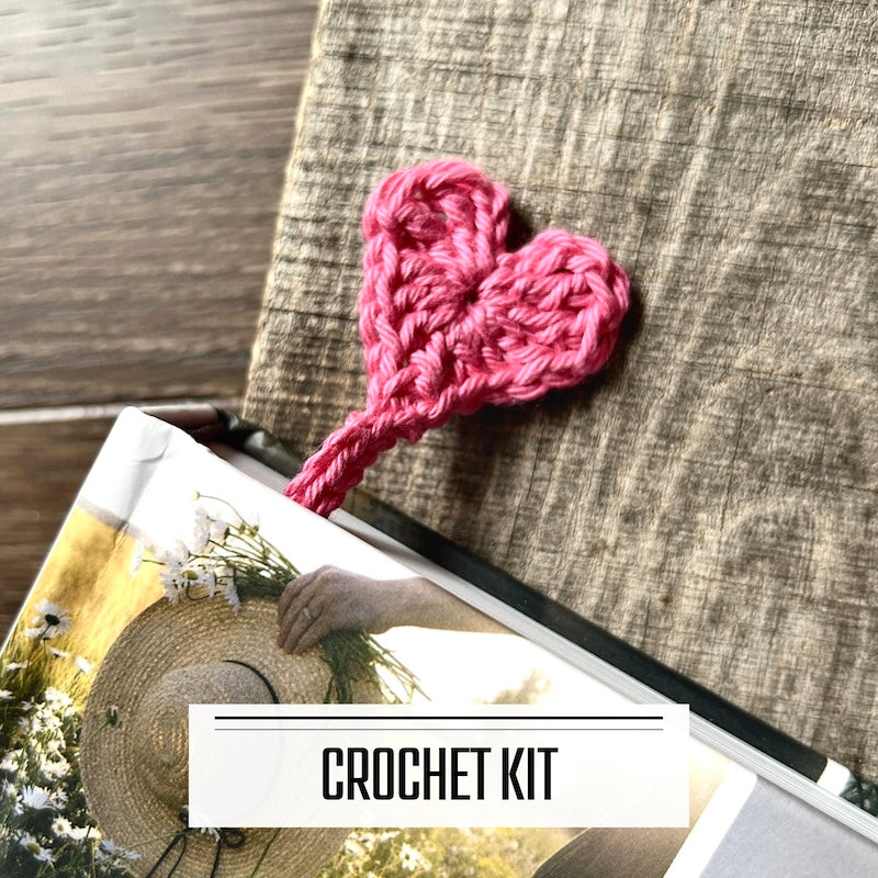 A DIY crochet kit for heart bookmarks. The finished bookmarks are sitting on a wooden board, peeking out from the top of a book.