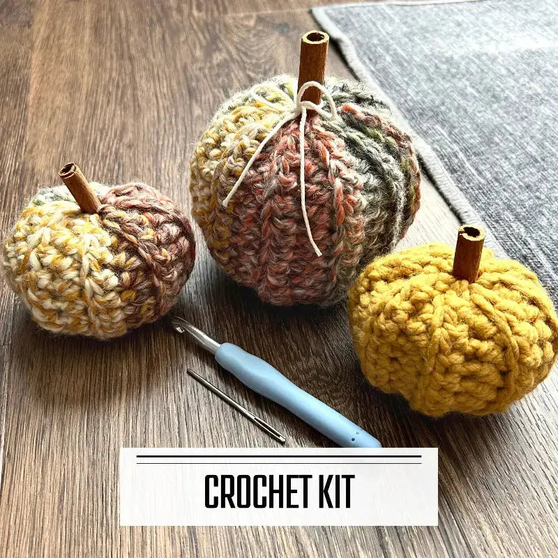 A crochet kit for a set of autumn pumpkins. The finished pumpkins are sitting on the floor with a crochet hook and tapestry needle in front of them.