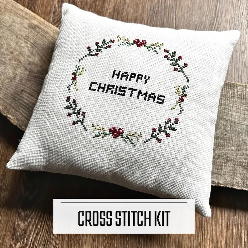 A DIY cross stitch kit for a Christmas throw pillow. The finished pillow is sitting on a wooden board.