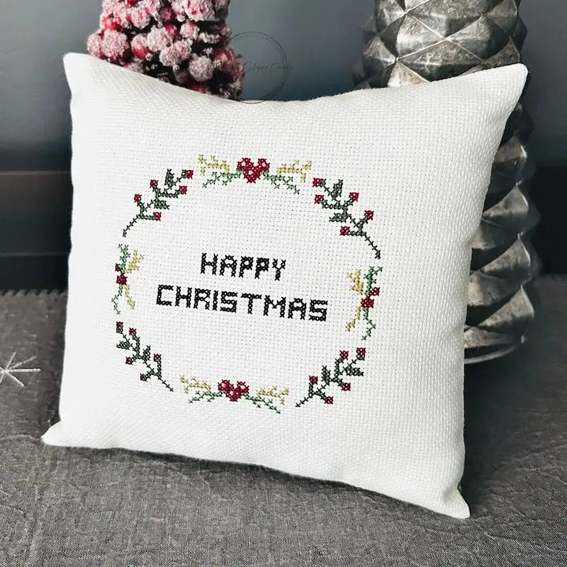 The finished pillow, made from this cross stitch pattern, is sitting on a table against 2 Christmas trees.