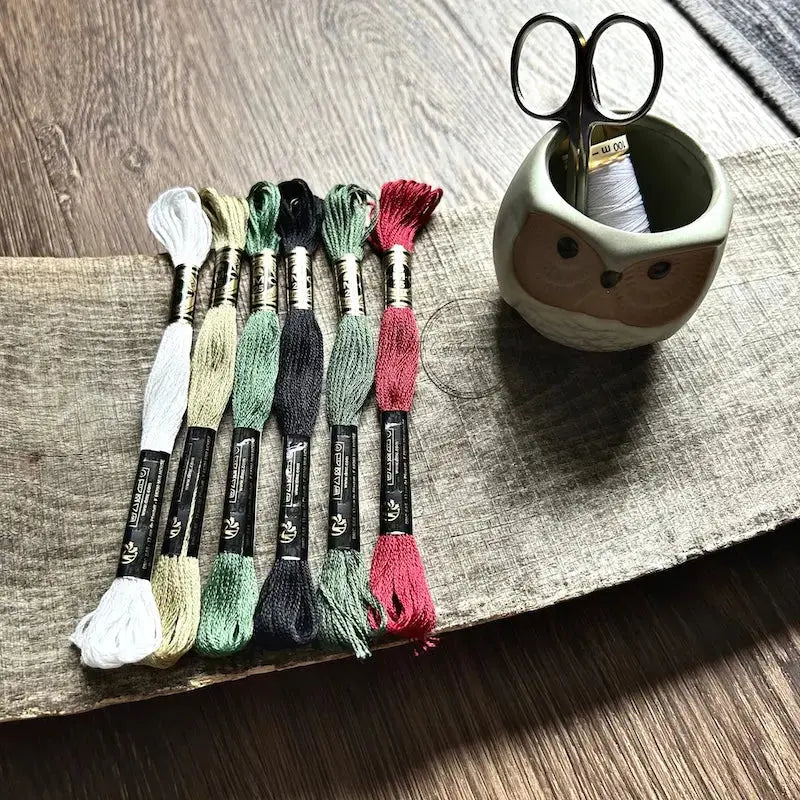 DMC floss, that would be used in this cross stitch pattern, is sitting on a wooden board with scissors and a spool of thread.