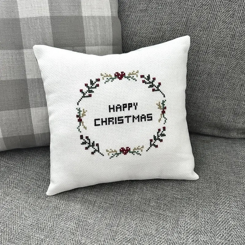 The finished pillow, made from this DIY cross stitch kit is sitting on a couch against a cushion.