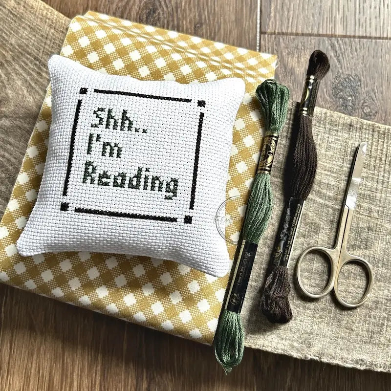 The finished pillow, made from this cross stitch kit, is sitting on a board with all of the supplies.