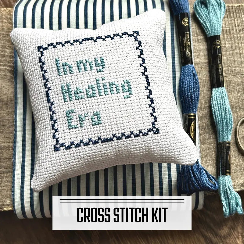 A DIY cross stitch kit for a mini pillow that can be used as a pin cushion. The finished pillow is sitting on a wooden board.
