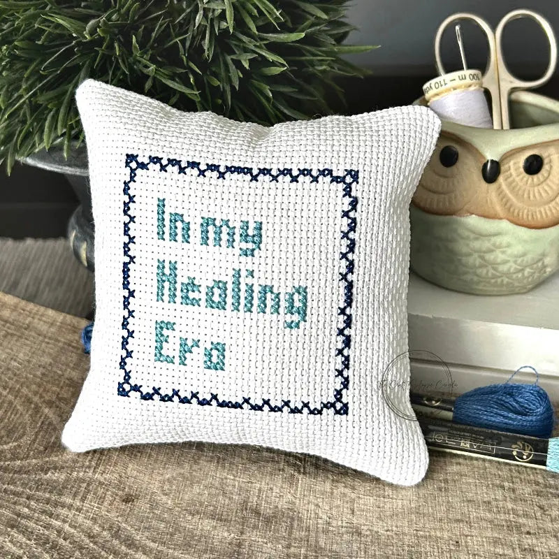The finished pillow, made from this cross stitch kit, is sitting on a wooden board.
