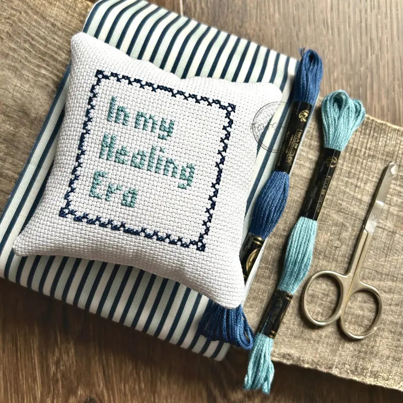The supplies included in your cross stitch kit are sitting on a wooden board.