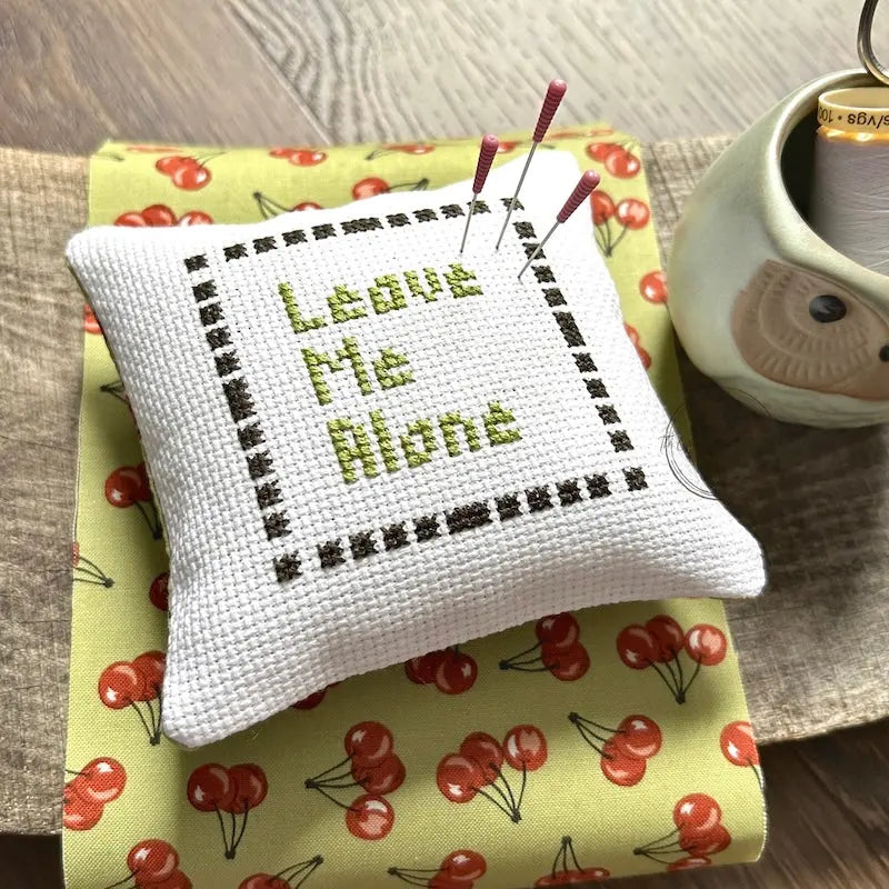 The finished pillow, made from this cross stitch pattern, is sitting on a wooden board with pins sticking out.