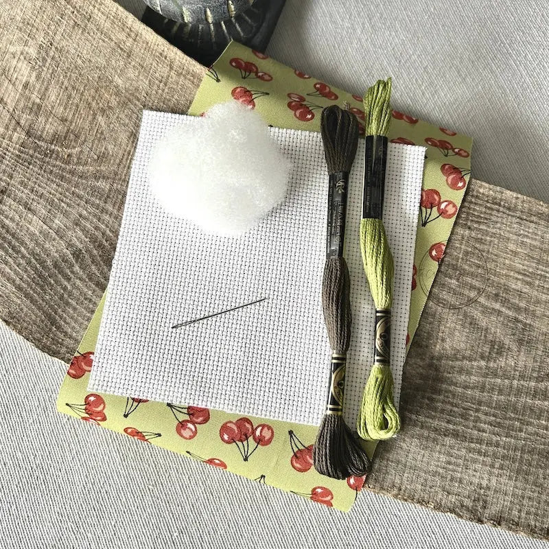 The supplies needed to make this cross stitch mini pillow are sitting on a wooden board.