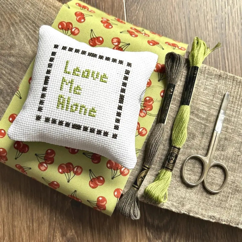 The finished pillow, made from this cross stitch kit, is sitting on a board with all of the supplies needed.