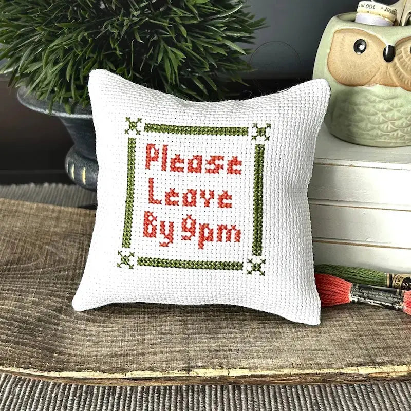The finished pillow, made from this cross stitch kit, is sitting on a wooden board.