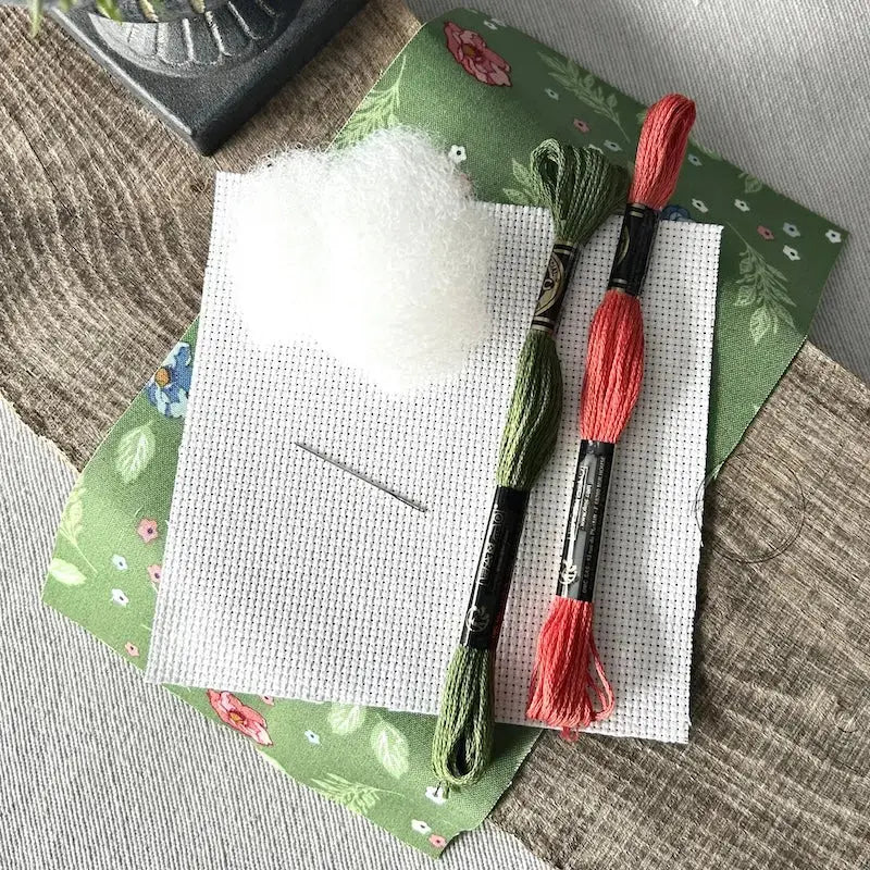 The supplies needed to make this cross stitch mini pillow are sitting on a wooden board.