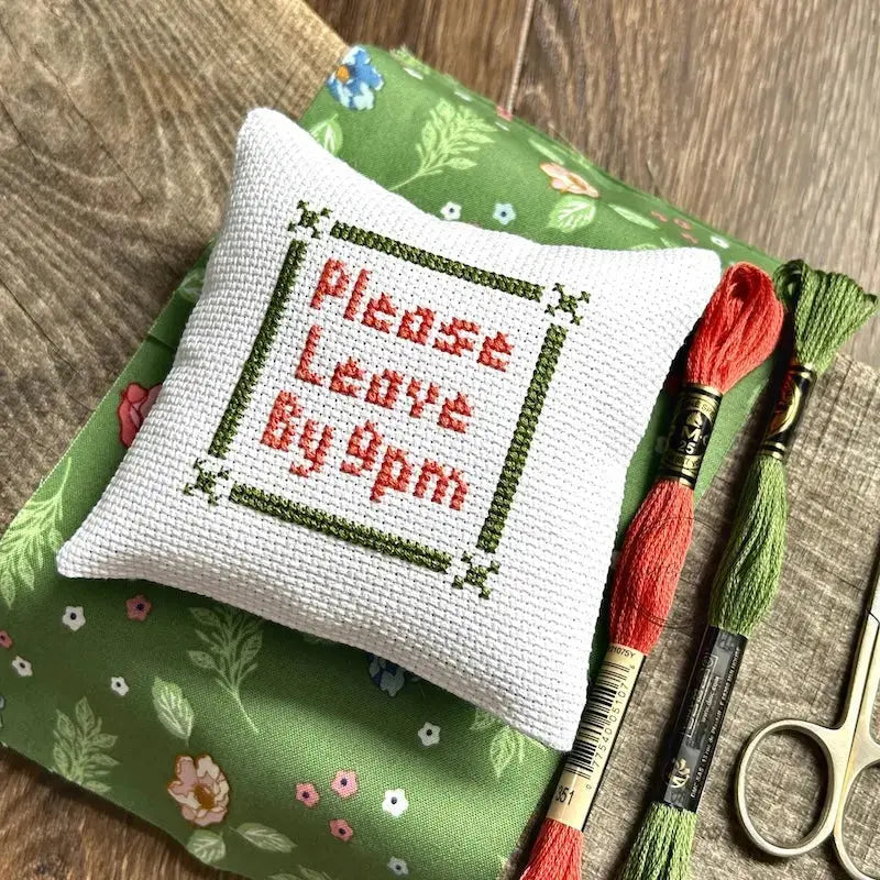The finished pillow, made from this cross stitch kit, is sitting on a board with all of the supplies needed.