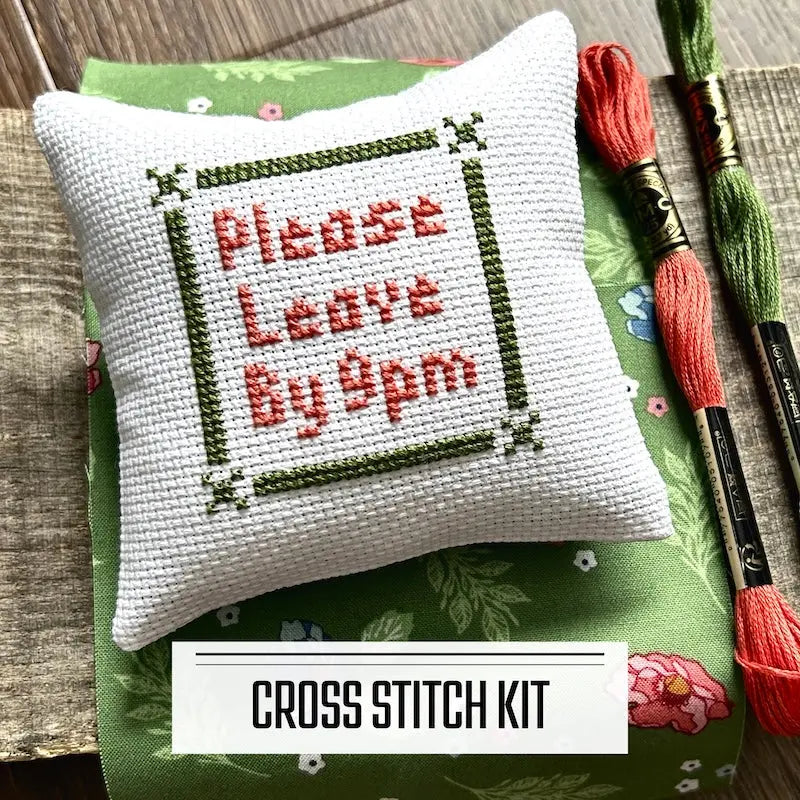 A DIY cross stitch kit for a mini pillow that can be used as a pin cushion. The finished pillow is sitting on a wooden board.