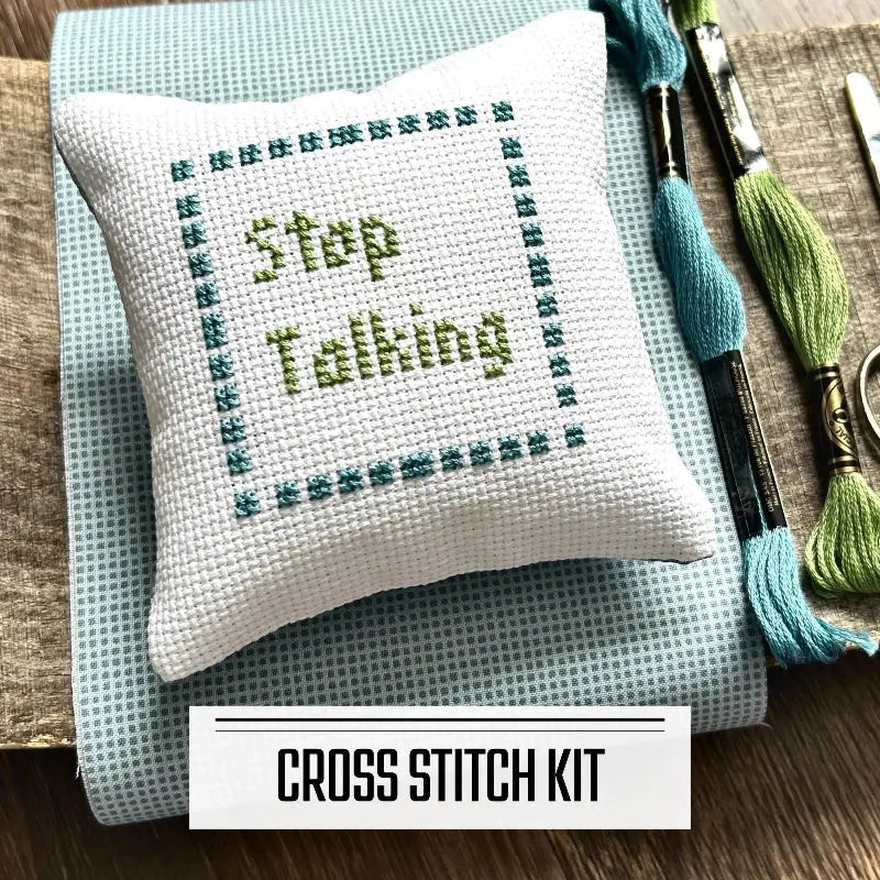 A DIY cross stitch kit for a mini pillow that can be used as a pin cushion. The finished pillow is sitting on a wooden board.