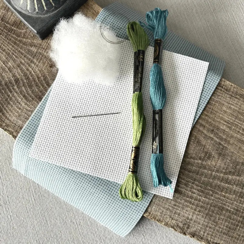 The supplies needed to make this cross stitch mini pillow are sitting on a wooden board.